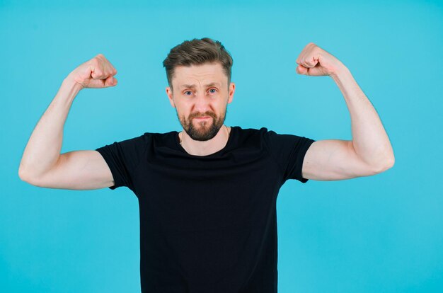 Der junge Mann hebt seine Muskeln auf blauem Hintergrund