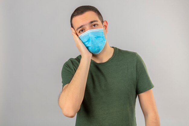 Der junge Mann sieht krank aus und trägt eine Gesichtsmaske gegen das Corona-Virus. Covid-9 berührt seine Wange auf Weiß