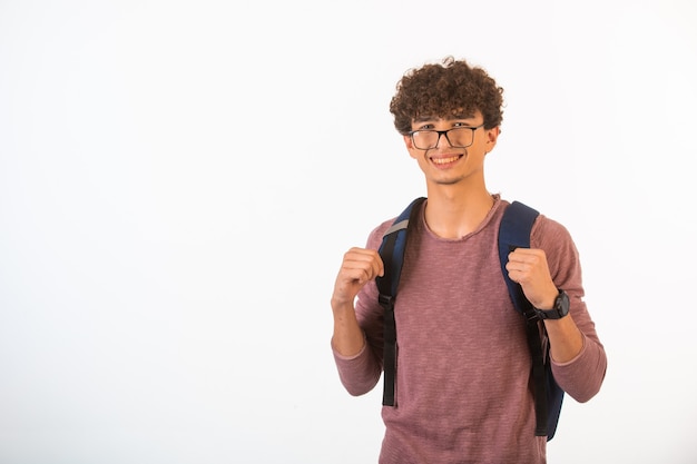 Der Junge mit dem lockigen Haar in der optischen Brille, der seinen Rucksack hält, ist zuversichtlich und motiviert, Vorderansicht.