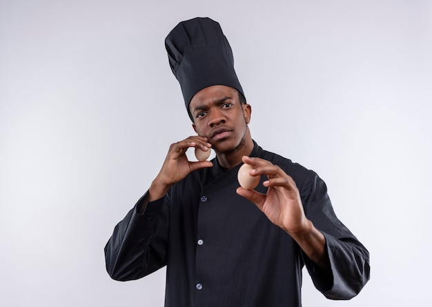 Der junge selbstbewusste afroamerikanische Koch in der Kochuniform hält Eier und gibt vor, isoliert auf weißem Hintergrund mit Kopienraum zu verteidigen