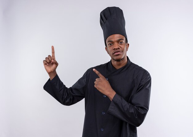 Der junge selbstbewusste afroamerikanische Koch in der Kochuniform zeigt mit beiden Händen lokalisiert auf weißem Hintergrund mit Kopienraum
