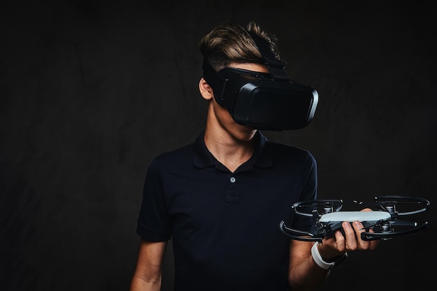 Der junge Teenager in einem schwarzen T-Shirt trägt eine Virtual-Reality-Brille und hält einen Quadrocopter. Auf dem dunklen Hintergrund isoliert.