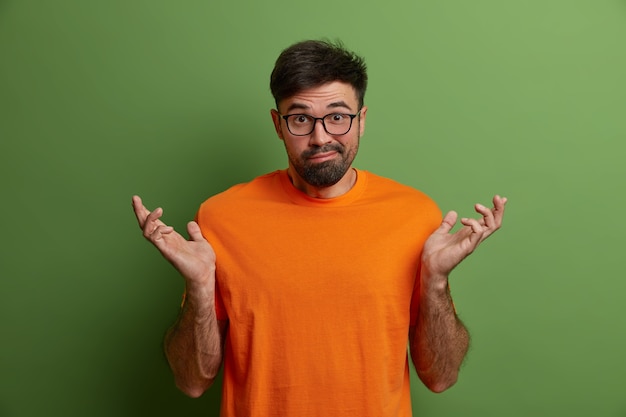Der kaukasische Mann breitet die Hände seitwärts aus, steht ahnungslos da, hat keine Ahnung, was passiert ist, ist verwirrt zu antworten, gekleidet in ein orangefarbenes T-Shirt, isoliert auf einer grünen Wand. Was ist los. Unentschlossener Freund