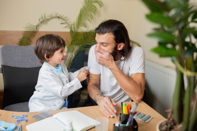 Der kleine Junge, der spielt, tut so, als ob ein Arzt einen Mann in einer komfortablen Arztpraxis untersucht