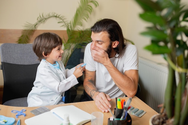 Der kleine Junge, der spielt, tut so, als ob ein Arzt einen Mann in einer komfortablen Arztpraxis untersucht