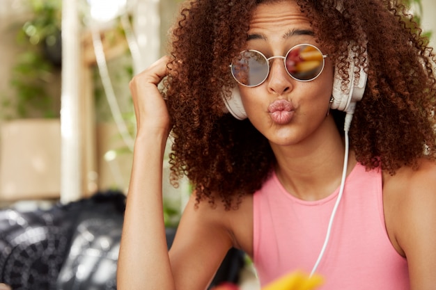 Der Kopfschuss einer angenehm aussehenden, stilvollen Frau in Schattierungen hat eine afrikanische Frisur, runde Lippen, einen lustigen Ausdruck, genießt Lieblingsmusik oder Audio in Kopfhörern auf der Radio-Website. Menschen- und Stilkonzept