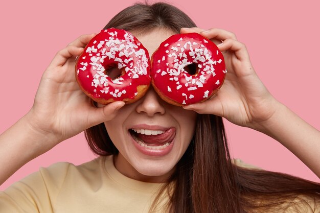 Der Kopfschuss einer lustigen jungen europäischen Frau bedeckt die Augen mit zwei glasierten Donuts, leckt die Lippen mit der Zunge und hat dunkles Haar