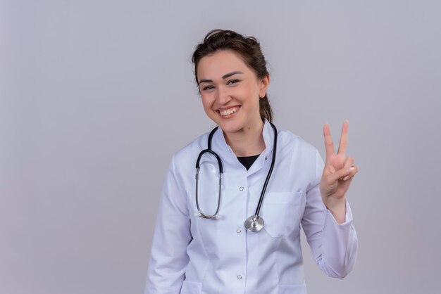 Der lächelnde junge Arzt, der das medizinische Kleid trägt, das Stethoskop trägt, zeigt Friedensgeste auf weißer Wand