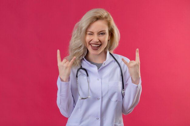 Der lächelnde junge Arzt, der ein Stethoskop im medizinischen Kleid trug, blinzelte und zeigte eine Ziegengeste auf einem roten Hintergrund