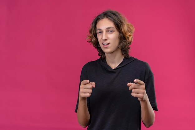 Der lächelnde Kerl mit den langen Haaren im schwarzen T-Shirt zeigt auf der rosa Wand nach vorne