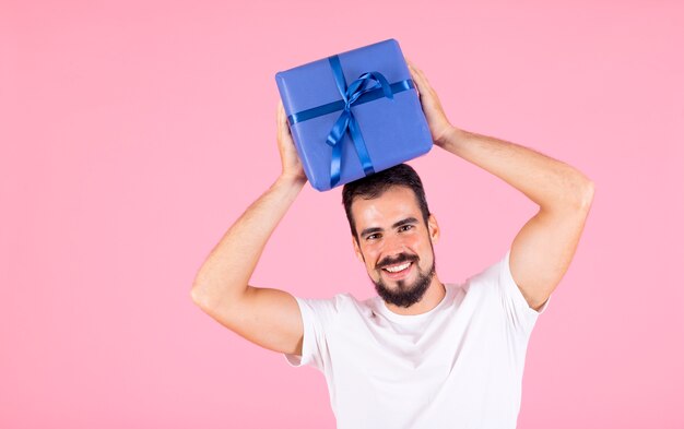 Der lächelnde Mann, der Blau hält, wickelte Geschenkbox über seinem Kopf gegen rosa Hintergrund ein