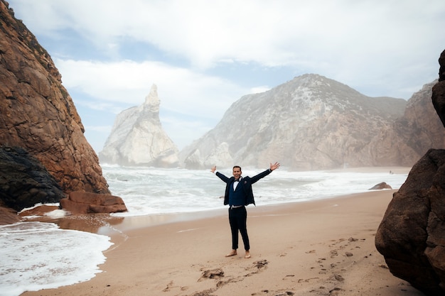 Der mann im anzug steht am strand zwischen den felsen und sieht glücklich aus