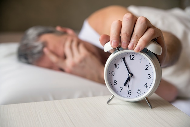 Der Mann liegt im Bett und schaltet am Morgen um 7 Uhr einen Wecker aus. Attraktiver Mann schläft in seinem Schlafzimmer. Verärgerter Mann, der durch einen Wecker in seinem Schlafzimmer geweckt wird