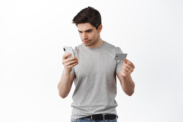 Der Mann schaut ernst auf den Smartphone-Bildschirm, während er online mit der Lieferung der Bestellung per Plastikkreditkarte im Internet über weißem Hintergrund einkauft