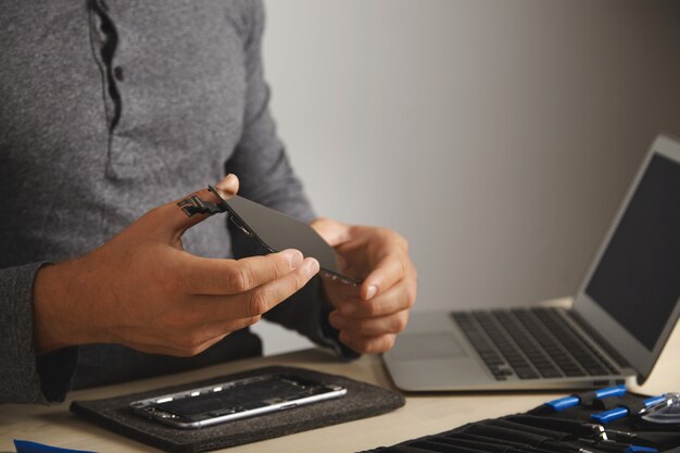 Der Meister hält einen neuen Bildschirm zum Ersetzen über dem zerlegten Smartphone in seinem Labor, einen Werkzeugsatz mit Instrumenten und einen Laptop vor sich auf einem weißen Tisch, Nahansicht