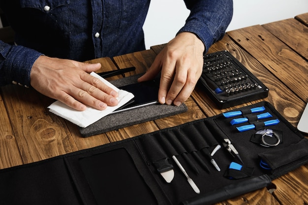 Der Meister reinigt das Mobiltelefon nach erfolgreicher Restaurierung mit einem weißen Tuch in der Nähe seiner professionellen Instrumente in einer Werkzeugtasche auf einem Holztisch