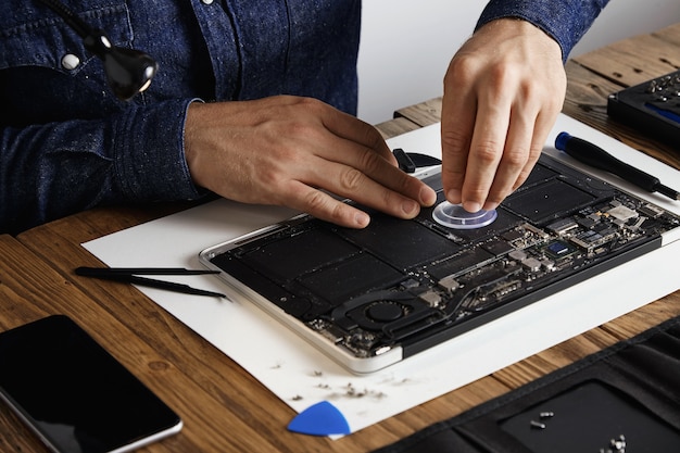 Der Meister verwendet einen kleinen Saugnapf, um die Batteriezellen eines kaputten Laptops auszutauschen, um sie in seinem Labor mit einem speziellen Werkzeugsatz auf einem Holztisch zu reparieren und zu reinigen