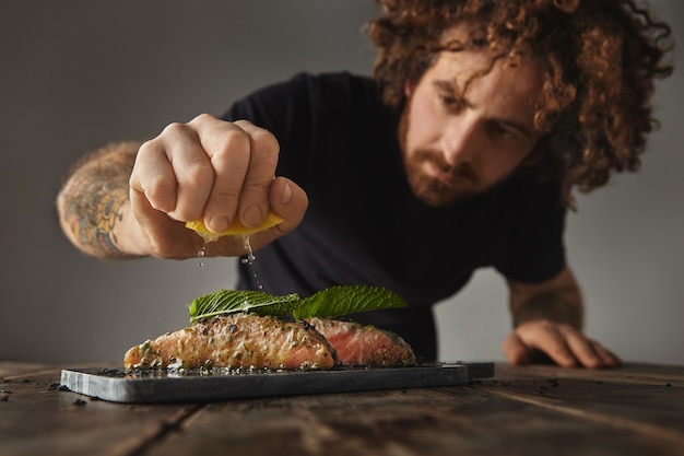 Der Mensch kocht eine gesunde Mahlzeit, drückt Limon auf zwei rohen Lachsstücken, die mit Minzblatt in Weißwein verziert sind, mit Gewürzen und Kräutern, die auf einem für den Grill vorbereiteten Marmordeck präsentiert werden