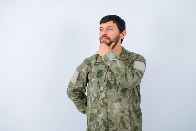 Der Militärmann denkt, indem er die Hand am Kinn hält und die andere Hand auf die Taille auf weißem Hintergrund legt
