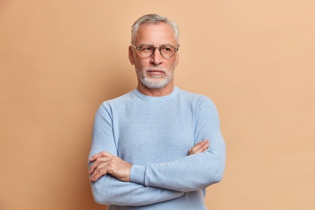 Der nachdenkliche alte grauhaarige Großvater hält die Arme verschränkt und schaut nachdenklich über etwas Wichtiges nach, das in einem lässigen Pullover gekleidet ist