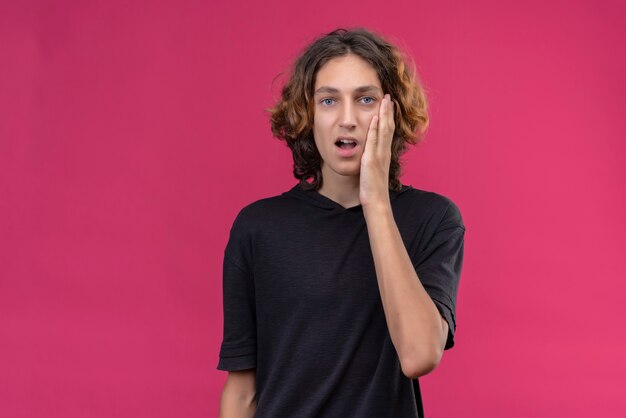 Der nachdenkliche Kerl mit den langen Haaren im schwarzen T-Shirt legte seine Hand auf die Wange an der rosa Wand