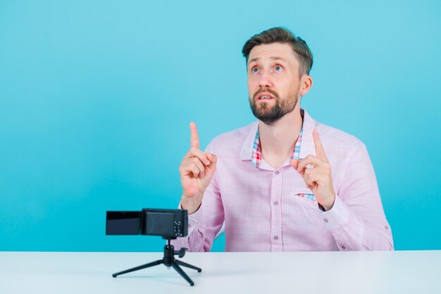 Der nachschlagende Blogger ma zeigt mit den Zeigefingern nach oben, indem er vor seiner Minikamera auf blauem Hintergrund sitzt