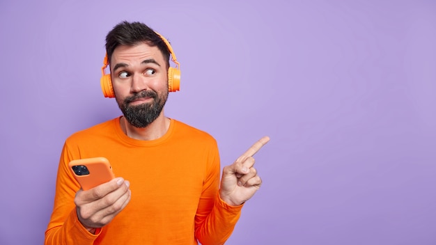 Der neugierige, gutaussehende Mann mit Bart zeigt etwas Interessantes an leeren Stellen an der lila Wand verwendet das Mobiltelefon zum Online-Chatten und das Hören von Musik trägt lässig gekleidete Kopfhörer