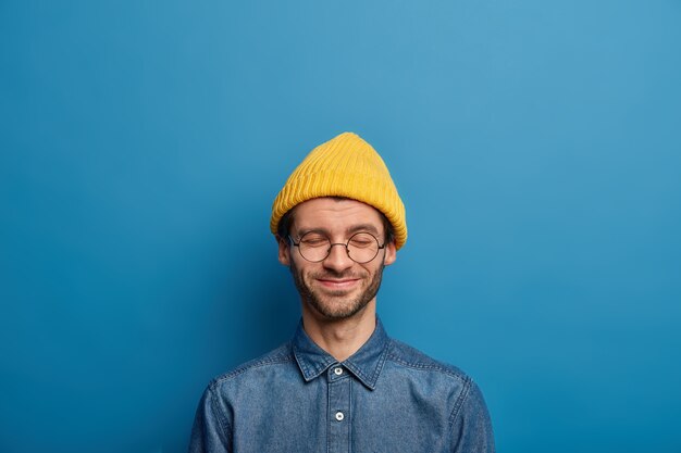 Der optimistische Caucaisan-Mann hält die Augen geschlossen, lächelt glücklich, trägt gelbe Kopfbedeckungen und ein Jeanshemd