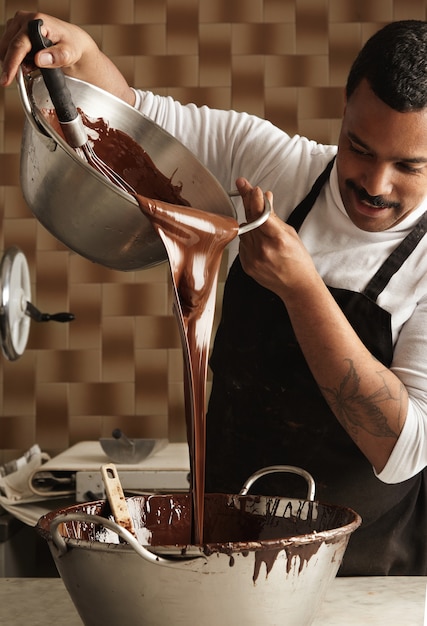 Der professionelle Chef des schwarzen Mannes gießt leckere geschmolzene Schokolade von einem großen Stahltopf in einen anderen, bevor er Schokoriegel herstellt