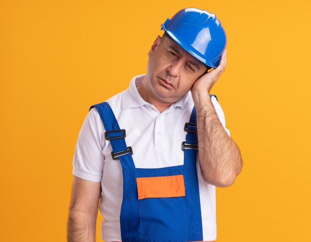 Der schmerzende erwachsene Baumeister in Uniform legt die Hand auf den Kopf, der auf der orangefarbenen Wand isoliert ist