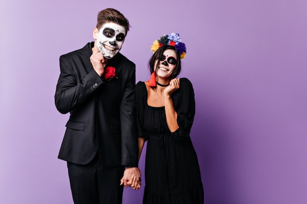 Der schuldige Freund und seine Freundin versuchen süß zu lächeln. Porträt der Frau mit hellen Accessoires und Mann im dunklen klassischen Anzug, der mit Make-up für Halloween aufwirft.