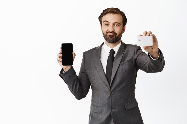 Der skeptische Geschäftsmann, der seine Kreditkarten-Smartphone-Bildschirm-Handyschnittstelle zeigt, mag etwas nicht, das über weißem Hintergrund steht