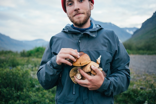 Der stolze Pflücker in einem traditionellen blauen Wollpullover mit Ornamenten steht auf einem Campingplatz in den Bergen und hält in den Armen einen Haufen köstlicher und biologischer Pilze