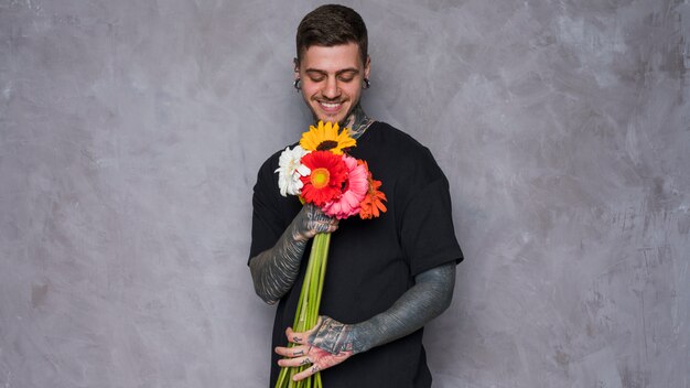 Der tätowierte junge Mann, der bunten Gerbera betrachtet, blüht in der Hand gegen grauen Hintergrund