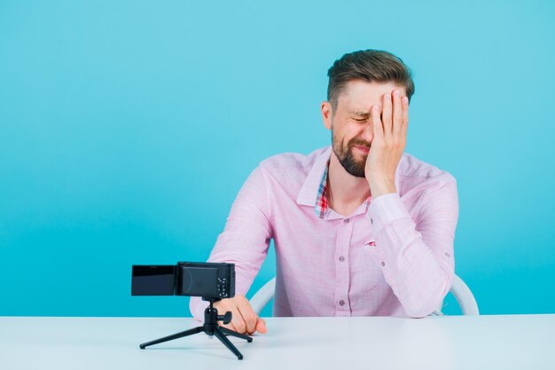Der traurige Blogger-Mann hält die Hand auf dem Gesicht, indem er vor seiner Mini-Kamera auf blauem Hintergrund sitzt