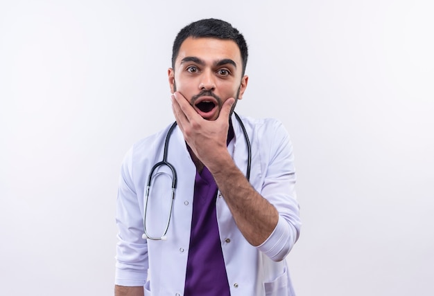Der überraschte junge männliche Arzt, der das medizinische Kleid des Stethoskops trägt, legte seine Hand auf Kinn auf isoliertem Weiß