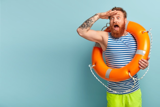 Der überraschte junge Urlauber mit roten Haaren und Bart kommt mit Sicherheitsausrüstung an den Strand, da er nicht schwimmen kann