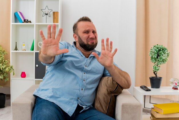 Der verängstigte erwachsene slawische Mann sitzt auf einem Sessel und streckt seine Hände aus und schaut in die Kamera im Wohnzimmer