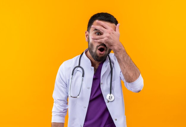Der verängstigte junge männliche Arzt, der das medizinische Kleid des Stethoskops trägt, legte seine Hand auf Auge auf lokalisierten gelben Hintergrund