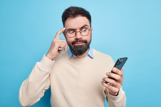 Der verlegene bärtige Mann, der sich auf das Smartphone konzentriert, hält den Finger auf den Tempel und versucht, sich auf die Informationen zu konzentrieren, die in ordentlichen Pullover-Posen gegen die blaue Wand gekleidet sind. Technologiekonzept