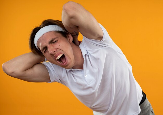 Der verletzte junge sportliche mann, der stirnband und armband trägt, packte den schmerzenden hals, der auf der orangefarbenen wand isoliert wurde