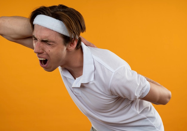 Der verletzte junge sportliche Mann, der Stirnband und Armband trägt, packte die schmerzende Taille, die auf der orangefarbenen Wand isoliert ist