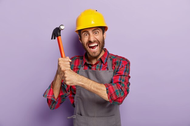 Der verzweifelte männliche Arbeiter oder Handwerker hält den Hammer mit beiden Händen, hat einen empörten Gesichtsausdruck, ist bereit zum Reparieren oder Bauen, trägt einen Schutzhelm, arbeitet auf der Baustelle, steht drinnen.