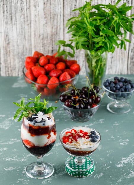 Dessert in Vase und Becher mit Erdbeeren, Blaubeeren, Minze, Kirschen High Angle View auf Gips und grungy Oberfläche