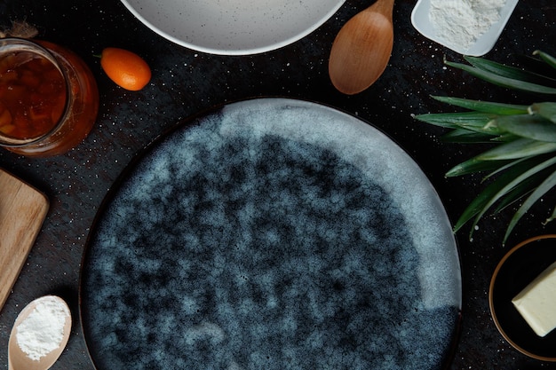 Detailansicht des leeren Tellers mit Marmelade, Kumquats, Mehl, Butter, Ananasblätter, Löffel auf schwarzem Hintergrund