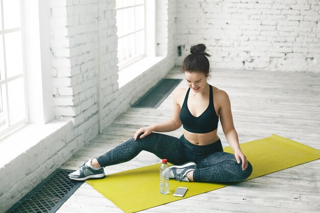 Diät- und Fitnesskonzept. Freudiges süßes Mädchen in der trendigen Sportbekleidung, die am Fenster sitzt und fröhlich lächelt und den Bildschirm des Smartphones betrachtet, das auf grüner Matte vor ihr liegt und Textnachricht liest