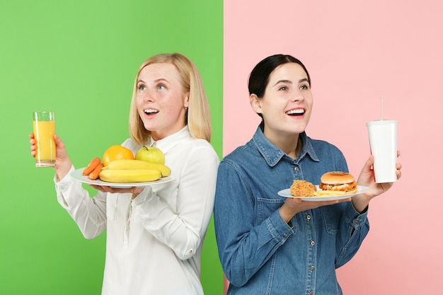 Diätkonzept. Gesundes nützliches Essen. Schöne junge Frauen, die im Studio zwischen Früchten und ungesundem Fast Food wählen. Menschliche Emotionen und Vergleichskonzepte