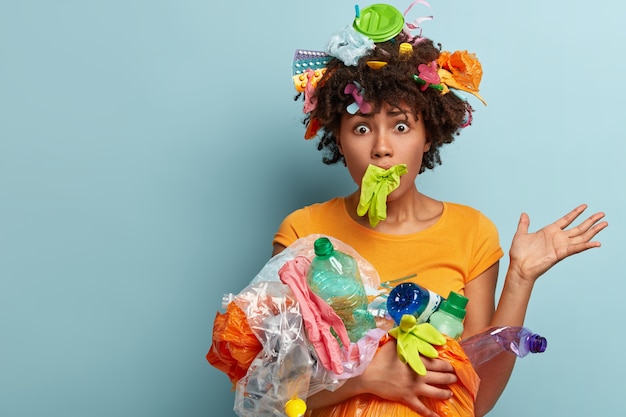 Die ängstliche, frustrierte afroamerikanische Frau ist mit Plastik überladen, hat den Mund mit einem Gummihandschuh festgeklemmt, hat Augen herausgesprungen, ist besorgt über die Naturverschmutzung, hilft bei der Reinigung der Umwelt und bietet Platz für Text
