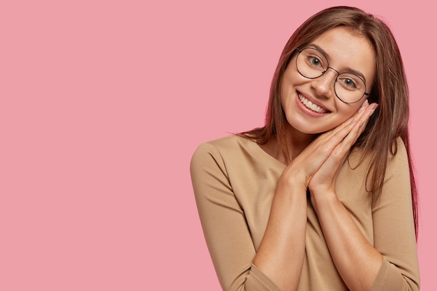 Die Aufnahme einer zufriedenen, zufriedenen jungen Frau hält die Handflächen zusammen, als ob sie ein Nickerchen machen würde, hat einen positiven Ausdruck, eine gesunde, saubere Haut, dunkles Haar und steht an der rosa Wand mit einem Kopierraum auf der rechten Seite