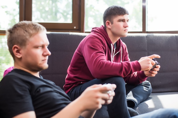 Die begeisterten Spieler. Zwei junge glückliche Männer, die Videospiele spielen, während sie auf Sofa sitzen